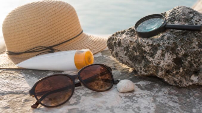 ein strohhut eine sonnencremeflasche eine sonnenbrille eine muschel und eine lupe liegen auf einer steinfläche in der nähe des wassers
