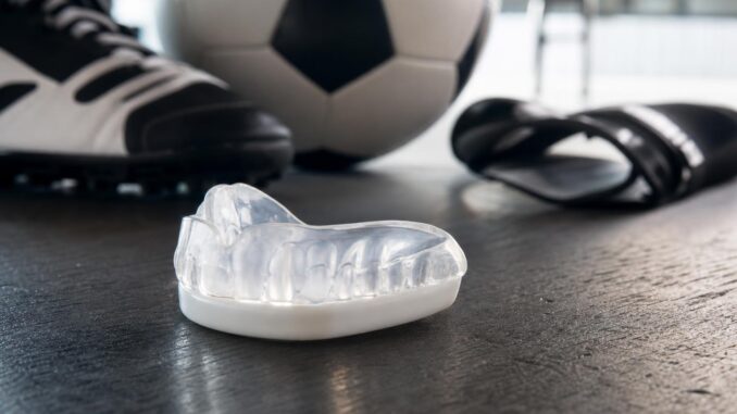 ein sportmundschutz liegt in einem fitnessstudio oder einer umkleidekabine auf dem boden neben einem fußballschuh einem fußball und einer slide sandale
